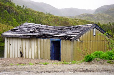 Arka planda çimenlerle kaplı tepeleri olan sarkık çatılı bir baraka.