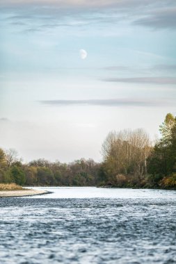 Gökyüzünde bulutlar olan ay ve yerde akan Mura nehri..