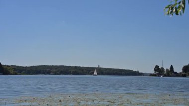 Berrak mavi gökyüzünün altında ormanla çevrili bir yelkenli ile huzurlu göl manzarası. Seyahat promosyonları, göl turizmi ve Avrupa doğa kaçamakları için ideal..