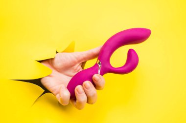 Sarı kağıt arka plandan sarkan mor vibratörlü kadın eli. Boşluğu kopyala