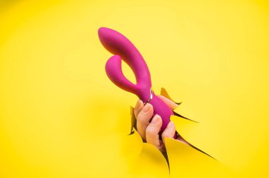 Sarı kağıt arka plandan sarkan mor vibratörlü kadın eli. Boşluğu kopyala