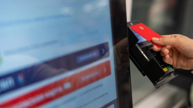 Bu resim, bir otomatik ödeme terminaline yaklaşırken kırmızı bir kredi kartı tutan bir eli yakalar.