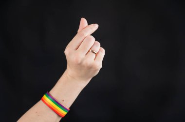 Alyansı ve gökkuşağı bileziği olan kadın siyah arka planda kalp işareti gösteriyor.