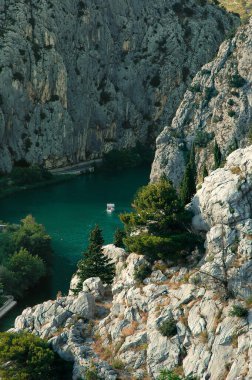 Güneşli bir günde, yüksek, kayalık dağlar arasında denizin ağzında yelken açan teknenin manzarası - hepsi yukarıdan görülür (Hırvatistan 'ın Omis kentinde çekilmiştir).)