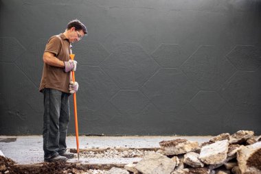 Eldivenli ve elinde turuncu levyeyle duran bir adam eldiven ve güvenlik gözlükleriyle inşaatta çalışıyor.