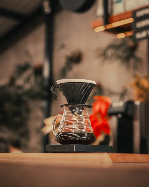 a glass carafe with coffee brewing, topped with a black coffee dripper, on a wooden surface