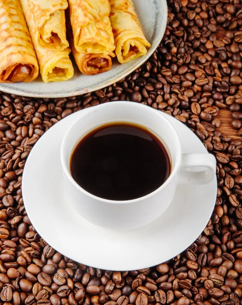 side view of a cup of coffee with wafer roll filled with condensed milk on a plate on coffee beans background