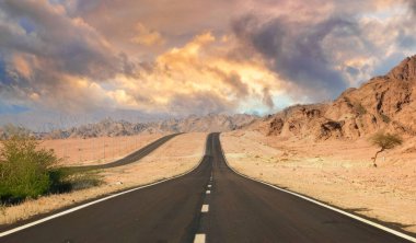 A wellmaintained twolane road leads through a desert near Dubai, flanked by rugged mountains under a vibrant sunrise or sunset sky, evoking solitude and adventure. clipart