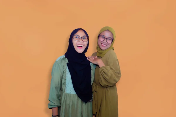 two muslim women wearing hijab standing and looking at camera over yellow background
