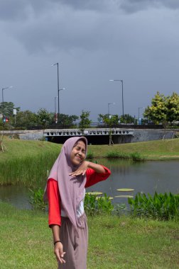 Pembe başörtüsü ve canlı giysileri olan bir kadın nehir kenarında durur, kolları ardına kadar açık, yaşama sevincini ve minnettarlığını ifade eder..