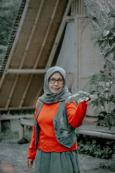 Peçe takan, kırmızı gömlek giyen bir kadın ağaç dallarını tutarken kameraya bakar.