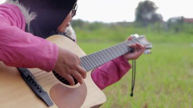 Açık hava ortamında akustik gitar çalan tesettür ve pembe ceket giyen biri. Ellere ve enstrümanlara odaklanmış, müziğin ve doğanın güzelliğini yakalıyor.