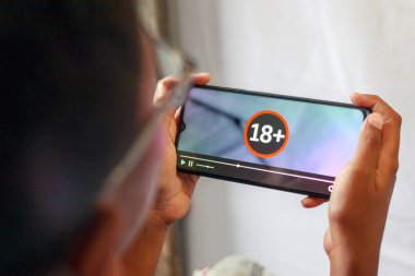Akıllı telefon ekranında yaş sınırlandırılmış içeriği izleyen bir kişi kapat