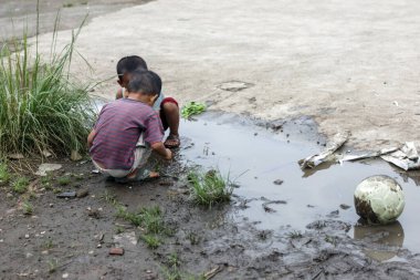 İki çocuk eski bir futbol topunun yanında çamurlu bir su birikintisinde oynuyor.