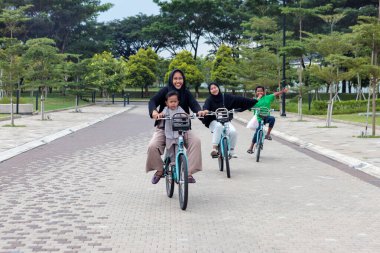 Neşeli bir aile, güzel yeşil bir parkta birlikte bisiklet sürüyor. Ağaçlar ve temiz havayla çevrili asfalt bir yolda açık hava aktivitelerinin tadını çıkarıyor.