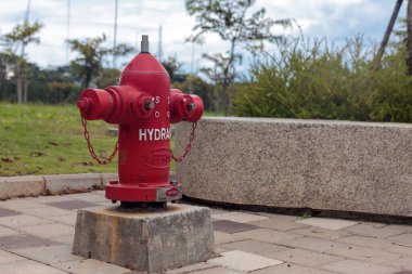 Güvenlik zincirleri olan kırmızı bir yangın musluğu halka açık bir parktaki beton bir üsse monte edilir. Bu temel itfaiye ekipmanları kaldırım ve yeşilliklerle çevrili, acil durum hazırlığını sağlıyorlar.