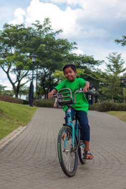 Yeşil gömlek giyen neşeli genç bir çocuk yemyeşil bir şehir parkında mavi bir bisiklete biniyor. Sahne, güneşli bir günde neşe, açık hava aktivitesi ve çocukluk macerasını yakalar.