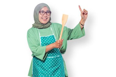 Neşeli Müslüman kadın önlük takıyor, elinde ahşap bir spatula tutarken gülümsüyor, ev yemeklerini, mutfak tutkusunu ve neşeli bir mutfak atmosferini temsil ediyor.