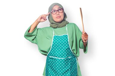 Ahşap bir spatula tutan tesettürlü ve önlüklü Müslüman bir kadın yemek pişirme fikirleri düşünüyor.