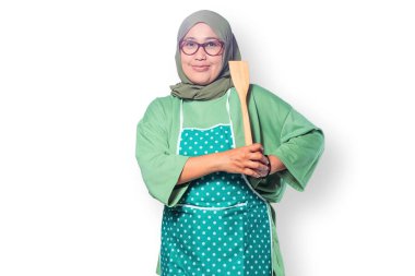 Mutfakta tahta bir spatula tutan tesettürlü ve önlüklü kendinden emin Müslüman bir kadın.