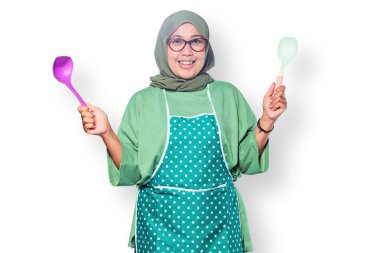 Mutfakta tesettür ve önlük takan mutlu Müslüman bir kadın yemek pişirme aletleri tutuyor.
