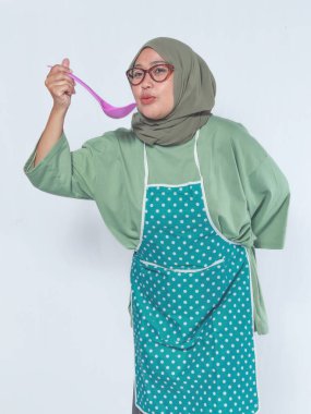 Müslüman kadın önlük takıyor kepçeyle yemek tadıyor, ev yemeklerini ve mutfak hazırlıklarını temsil ediyor.