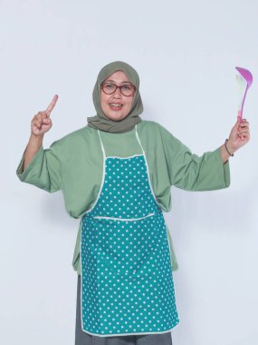 Önlüklü gülümseyen Müslüman kadın bir kepçeyle yemek pişirmekten zevk alıyor.