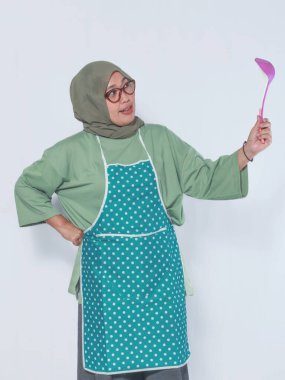 Önlüklü Müslüman bir kadın tuhaf bir ifadeyle kepçe tutuyor. Yemek ve yemek teması için mükemmel.