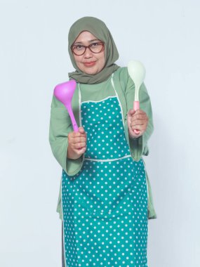 Önlük takmış, elinde kepçelerle gülümseyen Müslüman bir kadın.