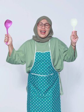 Önlük takan neşeli Müslüman kadın, elinde mutfak gereçleriyle mutlu mesut