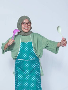 Mutfak gereçleri taşıyan önlüklü mutlu Müslüman kadın, yemek yapmaya hazır.