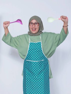 Neşeli Müslüman kadın şef önlük takıyor, mutfak gereçlerini tutuyor ve heyecanını dile getiriyor.