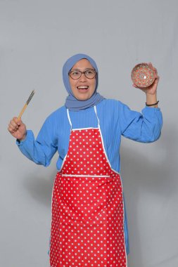 Mutfakta yemek pişirirken elinde yemek çubukları ve dekoratif bir kâse tutan neşeli Müslüman kadın.