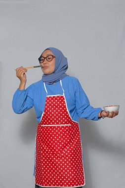 Önlüklü Müslüman bir kadın yemek çubuklarıyla yemek tadıyor, ev yemeği sahnesini resmediyor.