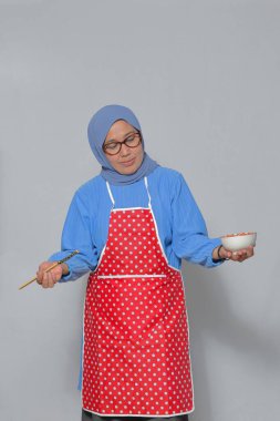 Önlüklü Müslüman bir kadın elinde yemek ve yemek çubuklarıyla yemek pişirme ve yemek temalarını temsil ediyor.