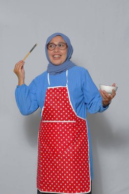 Önlüklü, yemek çubuğu ve kase tutan neşeli Müslüman kadın yemek pişirmekten zevk alıyor.