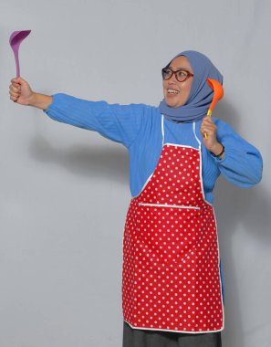 Önlük ve tesettüre bürünmüş enerjik Müslüman bir kadın mutfak aletlerini coşkuyla tutuyor.