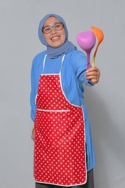 Önlüklü ve tesettürlü mutlu Müslüman bir kadın elinde neşeli bir gülümsemeyle mutfak aletleri tutuyor. Yemek pişirme ve mutfak temaları için mükemmel