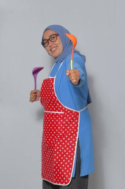 Önlük ve tesettüre bürünmüş kendinden emin Müslüman bir kadın, elinde neşeli bir ifadeyle mutfak aletleri tutuyor.