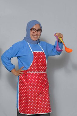 Mutfakta gülümserken elinde renkli kepçelerle önlük ve tesettür takan mutlu Müslüman kadın. Yemek pişirme ve mutfak temaları için mükemmel