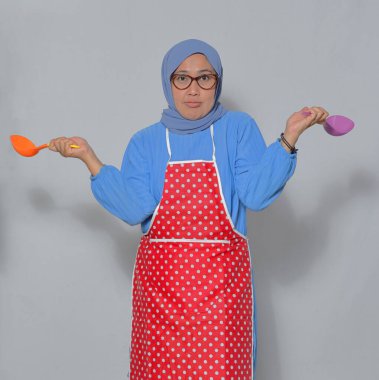 Mutfakta yemek pişirme aletleri tutan kafası karışmış Müslüman kadın, kendinden emin görünmüyor.