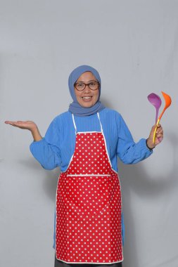 Mutlu Müslüman kadın önlük giyiyor, renkli mutfak aletleri tutuyor ve evde yemek pişirirken gülümsüyor.