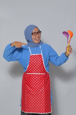 Heyecanlı Müslüman kadın önlük giyiyor, renkli mutfak aletleri tutuyor ve evde yemek hazırlarken gülümsüyor.