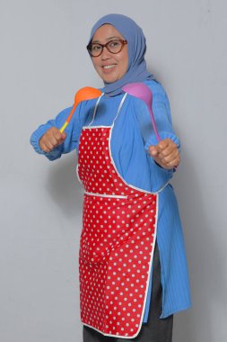 Mutlu Müslüman kadın önlük giyiyor, renkli mutfak aletleri tutuyor, evde yemek pişirirken gülümsüyor.