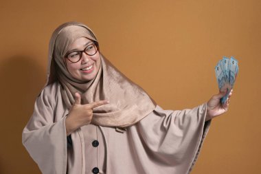 Endonezya parası taşıyan tesettürlü Müslüman bir kadın, mutlu bir ifadeyle finansal başarıyı ve zenginliği simgeliyor.