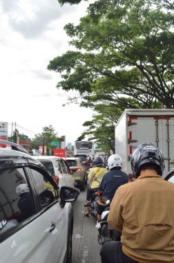 Kalabalık bir yolda trafik sıkışık. Ağaçlarla çevrili arabalar, motosikletler ve kamyonlar var. Bandung - Endonezya, 21 Ocak 2025