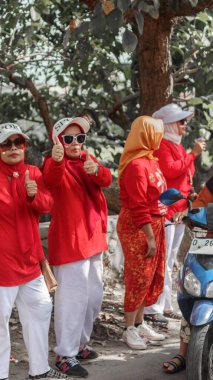 Bir grup yaşlı kadın, kırmızı kıyafetler ve beyaz pantolon giyip, dışarıda neşeli ifadeler, baş parmaklar ve güneş gözlükleriyle kutlama yapıyorlar, bir ağacın altında, Bandung - Endonezya, 17 Ağustos 2024