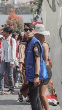 Mavi yelek ve güneş gözlüğü takan genç bir adam geçit töreninde kalabalığın arasında dikiliyor. Kırmızı ve beyaz giyinmiş insanlar olayı kutluyorlar. Bandung - Endonezya, 17 Ağustos 2024