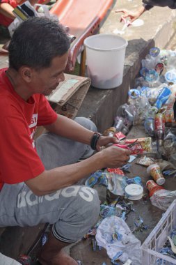 Bandung, Endonezya 29 Nisan 2025, Adam plastik atık yığınının yanında otururken geri dönüşüm için plastik ambalaj kesiyor