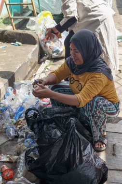 Bandung, Endonezya 29 Nisan 2025, Kadın temizlik sırasında plastik atığı siyah torbaya koyarken çömeliyor ve gülümsüyor